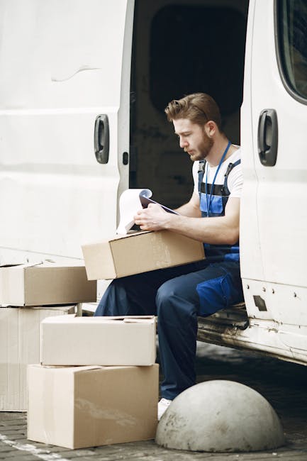 A male removal professional with light skin, short brown hair, and a beard is sitting on the doorstep of a white van, part of a house move. He is wearing a blue and white striped T-shirt and blue work overalls, focused on reading or reviewing a document in his hands. In front of him on the ground are several cardboard boxes of various sizes, some open and others sealed, likely used for packing household items. The van has its sliding side door open, revealing the dark interior space where additional packed boxes and moving equipment may be stored. The vehicle is parked close to a curb, with a concrete bollard nearby, indicating a typical urban or suburban setting for home relocation. The scene depicts a loading process in progress, with a focus on careful handling and packing logistics supported by [COMPANY_NAME], a professional removal service.