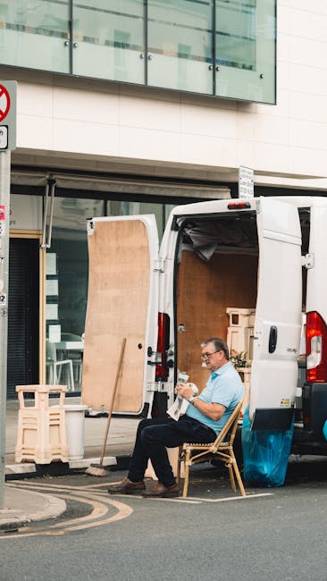 A male removal professional with light skin, short brown hair, and a beard is sitting on the doorstep of a white van, part of a house move. He is wearing a blue and white striped T-shirt and blue work overalls, focused on reading or reviewing a document in his hands. In front of him on the ground are several cardboard boxes of various sizes, some open and others sealed, likely used for packing household items. The van has its sliding side door open, revealing the dark interior space where additional packed boxes and moving equipment may be stored. The vehicle is parked close to a curb, with a concrete bollard nearby, indicating a typical urban or suburban setting for home relocation. The scene depicts a loading process in progress, with a focus on careful handling and packing logistics supported by [COMPANY_NAME], a professional removal service.