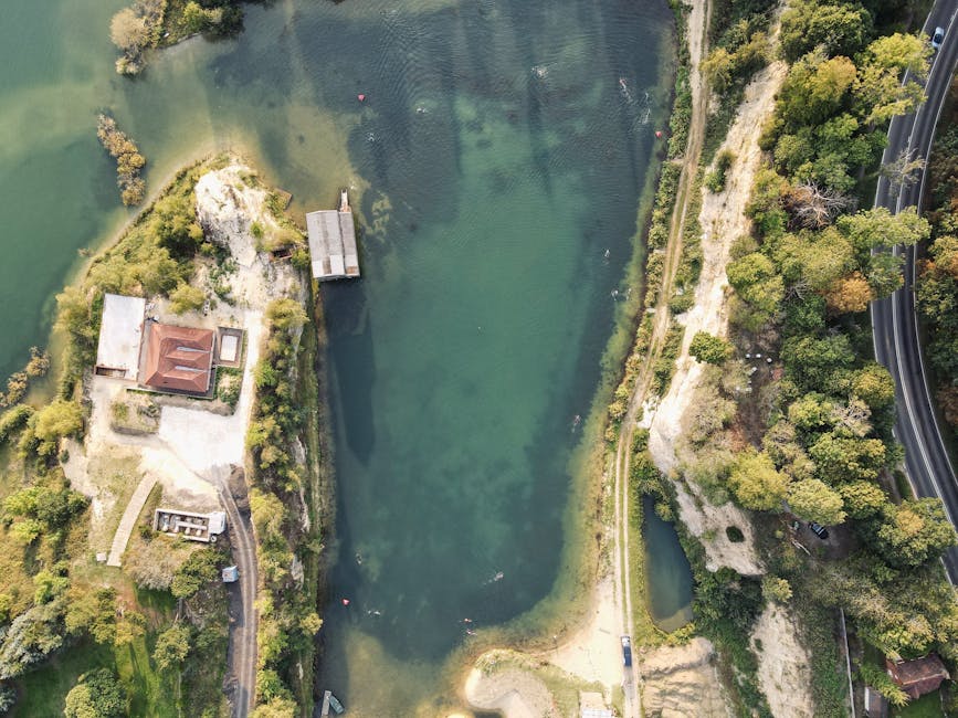 An aerial view of Bedfont Lakes in Feltham, showing a large body of water surrounded by greenery, trees, and pathways. To the left of the water, there is a small building with a reddish-brown roof and a nearby dock extending into the lake. Adjacent to this structure, a paved area contains several cardboard boxes and packing supplies, suggesting a packing and moving process. On the right side of the lake, a narrow dirt path runs along the water's edge, with dense trees and bushes lining the path and the surrounding area. To the far right, a section of a road with multiple lanes and a highway overpass is visible, with a line of vehicles moving along it. The scene captures a peaceful natural environment with ongoing activities related to house removals or furniture transport, supported by the presence of packing materials and the outdoor setting associated with a home relocation.