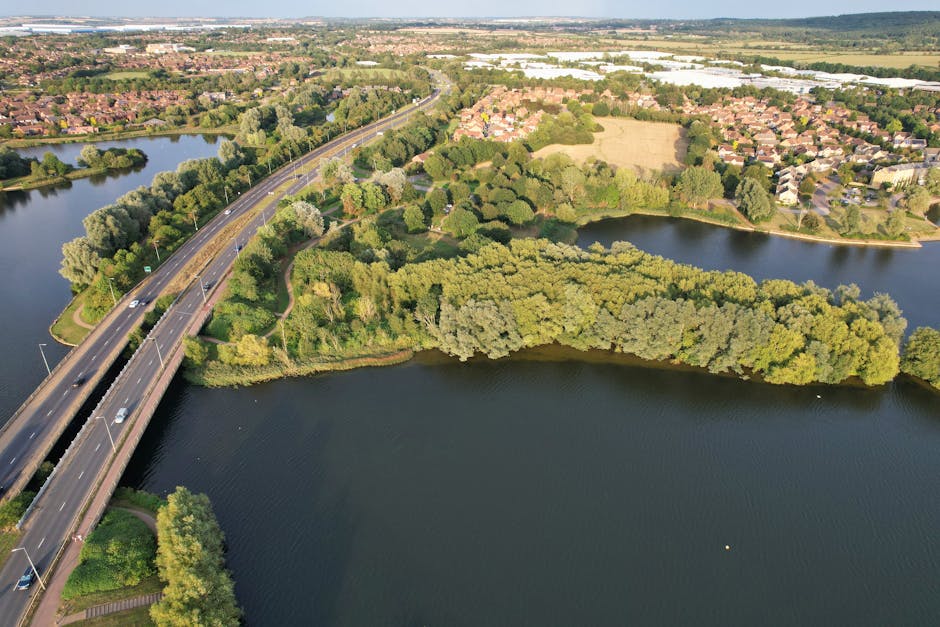 An aerial view showing a large body of water bordered by lush green trees and vegetation, with a bridge crossing over the water, supporting multiple lanes of vehicular traffic. On the far side, there is a residential area with numerous houses arranged in a suburban layout, interspersed with more greenery and open fields. The lighting indicates daytime with clear visibility of natural surroundings. This scene illustrates a peaceful suburban environment near Bedfont Lakes, Feltham, suitable for house removals and furniture transport, with the bridge and road network essential for efficient moving logistics, as managed by companies like Man with Van Feltham focusing on packing and moving services.