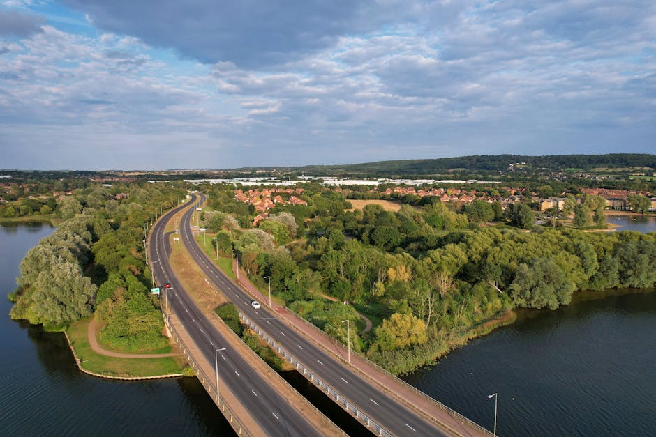 An aerial view of Bedfont Lakes in Feltham, showing a large body of water surrounded by greenery, trees, and pathways. To the left of the water, there is a small building with a reddish-brown roof and a nearby dock extending into the lake. Adjacent to this structure, a paved area contains several cardboard boxes and packing supplies, suggesting a packing and moving process. On the right side of the lake, a narrow dirt path runs along the water's edge, with dense trees and bushes lining the path and the surrounding area. To the far right, a section of a road with multiple lanes and a highway overpass is visible, with a line of vehicles moving along it. The scene captures a peaceful natural environment with ongoing activities related to house removals or furniture transport, supported by the presence of packing materials and the outdoor setting associated with a home relocation.