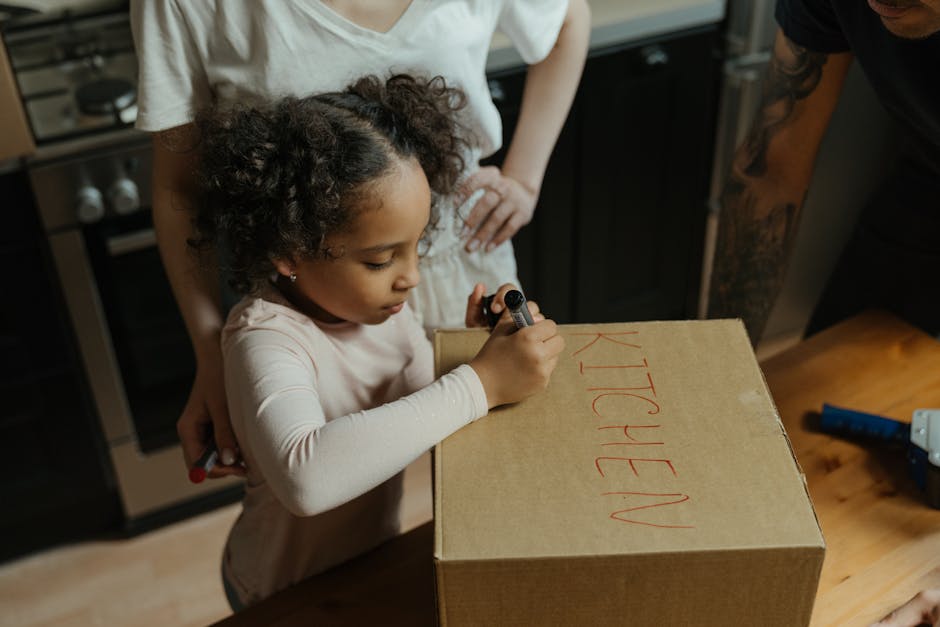 A young girl with curly hair wearing a long-sleeved beige shirt is standing at a wooden table inside a home, writing the word 'KITCHEN' in red marker on a plain cardboard box. An adult, partially visible, stands next to her, supporting her with one hand on her shoulder. The scene is set in a kitchen with dark cabinets, an oven, and a countertop in the background. On the table, there are some tools and packing materials, indicating a home relocation and furniture transport process. The image captures the packing stage of house removals, with children involved in labeling household items for an organized move. The lighting is warm and natural, emphasizing the detail and focus on the packing activity, which is part of the overall moving and packing preparations facilitated by companies like Man with Van Feltham.