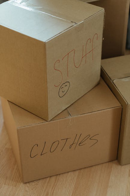 Close-up of three stacked cardboard moving boxes placed on a wooden floor inside a property. The top box has the word 'STUFF' handwritten in red marker along with a simple smiley face; the middle box has the word 'CLOTHES' written similarly, and the third box on the bottom is partially visible. The boxes are sealed with clear packing tape. The background shows additional boxes and packaging materials, indicating an ongoing home relocation process. The presence of handwritten labels suggests organized packing by [COMPANY_NAME], a professional removals service, supporting efficient furniture transport and logistics for homeowners preparing for their move, in line with the moving tips outlined for Feltham Centre flats near High Street.