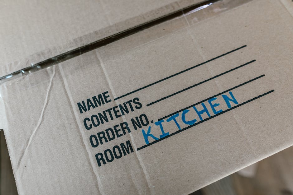 A woman sitting cross-legged on a wooden floor inside a property, surrounded by numerous cardboard boxes of various sizes, some sealed with red tape and others open showing packing materials. She is w
