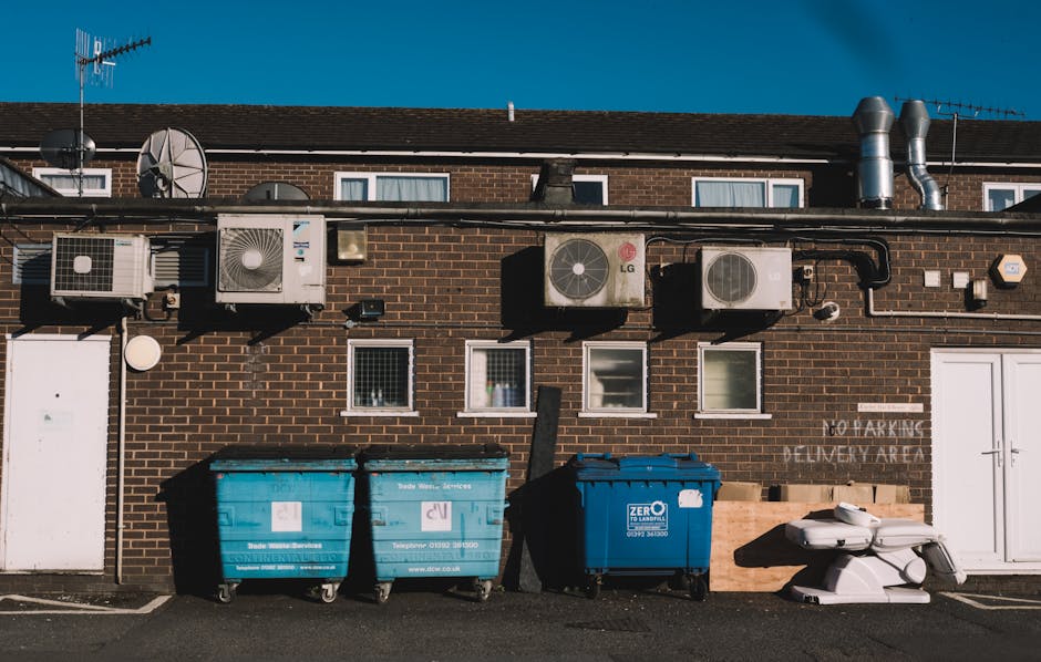 A row of three large waste collection bins, two blue and one green, positioned against the exterior brick wall of a residential building. The containers are situated on a paved surface directly outside a white double door and beneath a set of multiple air conditioning units and ventilation ducts attached to the building's upper wall. The background features a clear blue sky, and the area appears to be part of a driveway or designated waste disposal zone. A sign on the wall nearby indicates no parking or delivery activity. The scene is well-lit, with shadows cast by the bins and equipment, capturing the typical environment associated with home relocation logistics, waste management during moving processes, and the preparation involved in packing and disposal for a house move, as handled by Man with Van Feltham.