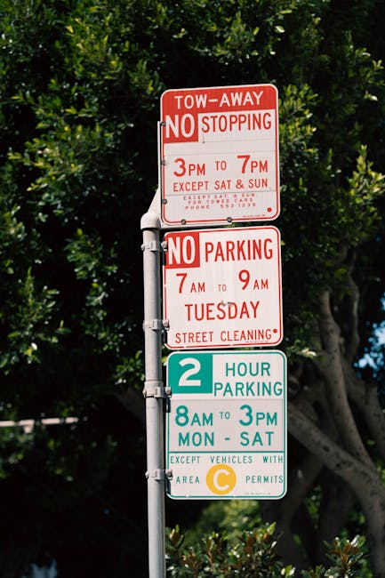 A vertical metal street pole with four traffic and parking restriction signs is situated on a pavement in Feltham, with dense green foliage and trees in the background. The top sign is red and white, indicating a tow-away zone with no stopping allowed from 3 pm to 7 pm, except on Saturdays and Sundays, designed to facilitate home relocation and furniture transport by managing street access during peak moving times. Below it, a similar red and white sign mandates no parking from 7 am to 9 am on Tuesdays for street cleaning purposes. The third sign is turquoise with white text, permitting two-hour parking from 8 am to 3 pm, Monday through Saturday, for vehicles with relevant area permits, supporting efficient loading and unloading during initial stages of packing and moving. The environment is well-lit, hinting at daytime, with the signs clearly visible for drivers and pedestrians, aiding in planning parking and street access during house removals or moving activities in Feltham. The presence of mature trees and dense foliage emphasizes a residential street setting, and some luggage or moving boxes might be temporarily staged nearby during relocation procedures, consistent with home moving logistics, supported by companies like Man with Van Feltham.