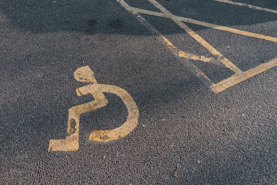 Close-up view of a designated disabled parking space on an asphalt surface, featuring a painted yellow wheelchair symbol that appears slightly worn and faded. The parking space is adjacent to a building entrance, with a portion of a curb and pavement visible in the background. Nearby are some yellow parking restriction lines, partially visible, indicating street access restrictions typical during house removals or furniture transport. The environment is well-lit, suggesting daytime conditions. This setting reflects typical moving day logistics, where careful parking permits or street access are necessary for efficient home relocation and furniture transport. The image is captured from an overhead angle, emphasizing the clear marking on the pavement, which is relevant to planning loading and unloading areas for professional removals services such as those provided by Man with Van Feltham.