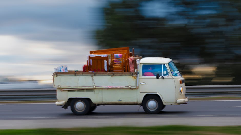 A vintage cream-colored van with a compact, rounded body and small windows is captured in motion along a road during daylight hours. The vehicle is loaded with household furniture and packed items, including wooden chairs, small tables, and various cardboard boxes contained within the open flatbed area. Some items are wrapped in plastic or fabric coverings to protect them during transit. The van's side mirror and front headlight are clearly visible, and the front windshield shows reflections from the surrounding environment. The background is blurred with streaks of greenery and sky, indicating the vehicle's movement. This image exemplifies the process of furniture transport and packing during home relocation, as carried out by moving professionals such as Man with Van Feltham, who specialise in removals and moving logistics, with attention to careful loading and vehicle safety.