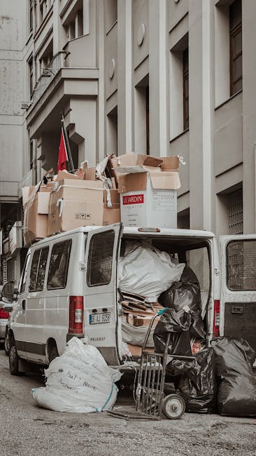 A white commercial van parked on a city street with its rear doors open, revealing a packed interior loaded with various moving supplies, including large black trash bags, cardboard boxes, and wrapped furniture. Several cardboard boxes are stacked on top of the van's roof, some open and filled with packing materials. A small hand trolley and a black plastic bag are positioned on the pavement beside the van, indicating ongoing loading or unloading activities. The van is situated next to a multi-storey building with light-colored stone or concrete exterior walls and several windows. This scene illustrates the process of home relocation and furniture transport, aligning with professional removals services by Man with Van Feltham, as part of packing and loading operations for a house move.