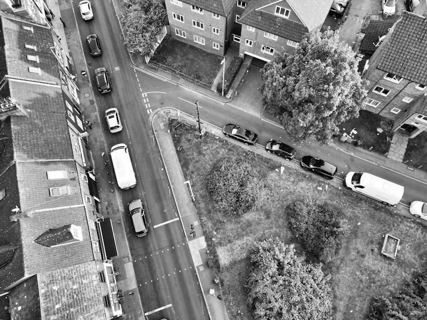A black and white aerial photograph showing a residential street with multiple parked cars along a narrow, curving road. On the left side, there are terraced houses with tiled roofs, chimneys, and front gardens, while on the right side, there is a small grassy area with several trees, bushes, and a paved pathway. A white van from Man with Van Feltham is parked on the pavement near the grassy area, adjacent to a doorway of one of the buildings. Nearby, multiple cardboard boxes, some wrapped in plastic, are visible on the pavement, indicating packing in progress for a house move. A hand truck or trolley is also present, being used to load or unload furniture or boxes. The scene suggests a home relocation process, with items being prepared for transport in the context of a move from Staines Road West to Feltham Station, involving professional removals from [COMPANY_NAME], with the environment lit evenly and the focus on loading or packing activities associated with furniture transport and packing and moving services.