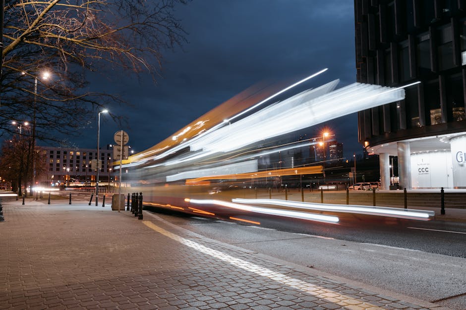 A long exposure photograph taken during evening or night showing light trails of a moving vehicle on a city street near a modern multi-storey building with glass windows. The street is lined with street lamps, with some trees visible on the left side. The pavement is made of brick-like tiles and a row of black metal bollards separates the sidewalk from the road. In the background, there are additional buildings and parked cars, with the scene illuminated by artificial lighting. This image captures the dynamic motion typical of urban moving and transportation, relevant in the context of house removals, furniture transport, and moving services provided by Man with Van Feltham, particularly during a home relocation process from Staines Road West to Feltham Station.