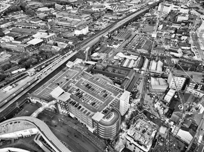 A black and white aerial photograph showing a residential street with multiple parked cars along a narrow, curving road. On the left side, there are terraced houses with tiled roofs, chimneys, and front gardens, while on the right side, there is a small grassy area with several trees, bushes, and a paved pathway. A white van from Man with Van Feltham is parked on the pavement near the grassy area, adjacent to a doorway of one of the buildings. Nearby, multiple cardboard boxes, some wrapped in plastic, are visible on the pavement, indicating packing in progress for a house move. A hand truck or trolley is also present, being used to load or unload furniture or boxes. The scene suggests a home relocation process, with items being prepared for transport in the context of a move from Staines Road West to Feltham Station, involving professional removals from [COMPANY_NAME], with the environment lit evenly and the focus on loading or packing activities associated with furniture transport and packing and moving services.