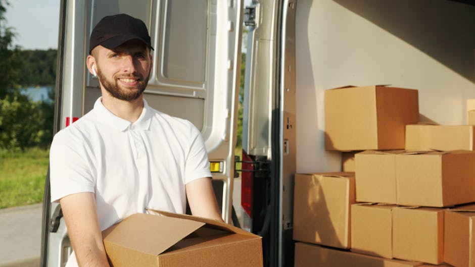 A man with a beard and wearing a white polo shirt and a black cap is standing next to an open moving van parked outside on a clear day. He is holding a brown cardboard box, preparing for a home reloca