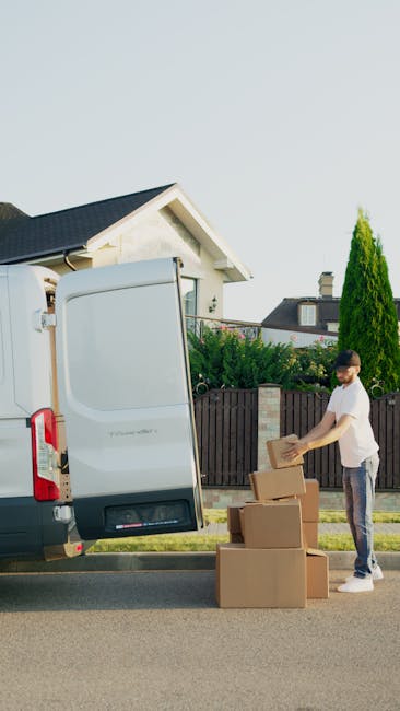 A man with a beard and wearing a white polo shirt and a black cap is standing next to an open moving van parked outside on a clear day. He is holding a brown cardboard box, preparing for a home reloca