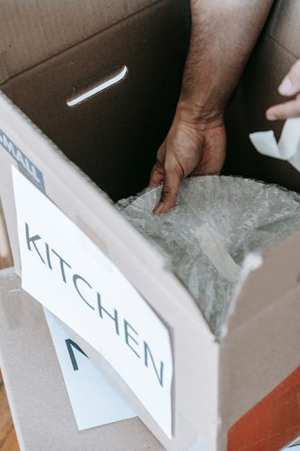 Close-up of a person’s hand placing bubble wrap inside an open cardboard box labeled 'KITCHEN', positioned on a flat surface within a home environment. The box is partially filled with protective pa