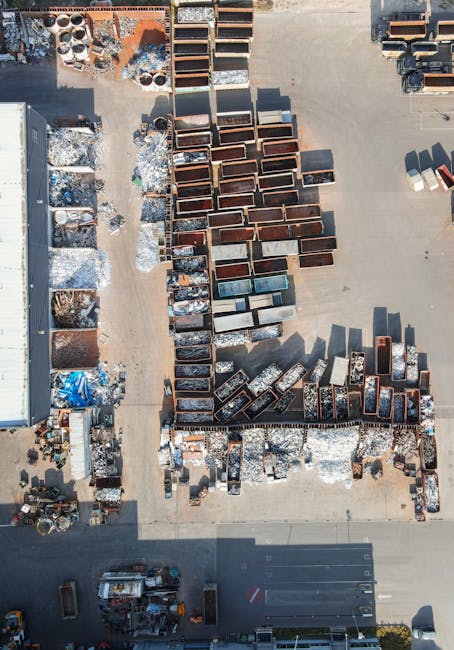 An aerial view of an outdoor area with a large number of discarded household items, including multiple metal and wooden furniture pieces such as cabinets, tables, and shelves arranged in rows on a concrete surface. Some items are piled or leaning against each other, with packaging materials like plastic wrap and cardboard boxes scattered nearby. A wheelbarrow and other yard tools are visible at the lower left corner, while a parked vehicle is at the bottom edge of the image. The background shows a building structure and additional debris, indicating a site for bulky waste disposal or clearance, relevant to house removals and relocation services provided by Man with Van Feltham during furniture transport and home relocation processes.